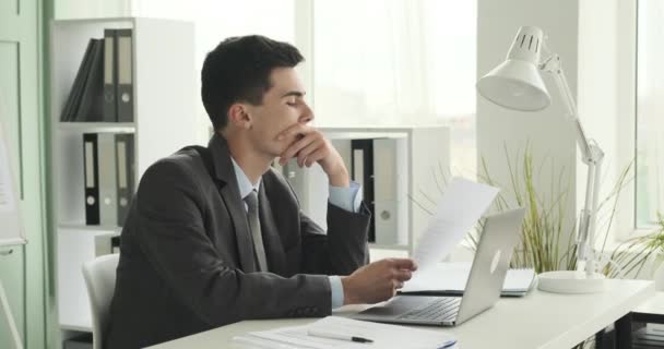 Młody i skupiony biały mężczyzna pracuje pilnie w swoim biurze. Siedzi przy biurku, zajęty swoimi zadaniami, z otwartym laptopem i rozłożonymi przed nim dokumentami.. - Materiał filmowy, wideo