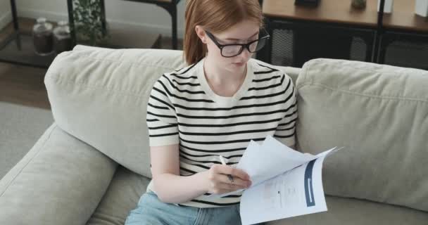 Una adolescente es capturada tomando notas en papeles mientras está sentada en un sofá. Con una expresión reflexiva, ella cuidadosamente escribe información importante o ideas. - Metraje, vídeo