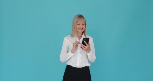 Optimistische vrouw wordt gelukkig zien surfen op haar telefoon op een serene blauwe achtergrond. Haar vrolijke uitdrukking en positieve houding suggereren een aangename en plezierige ervaring. - Video