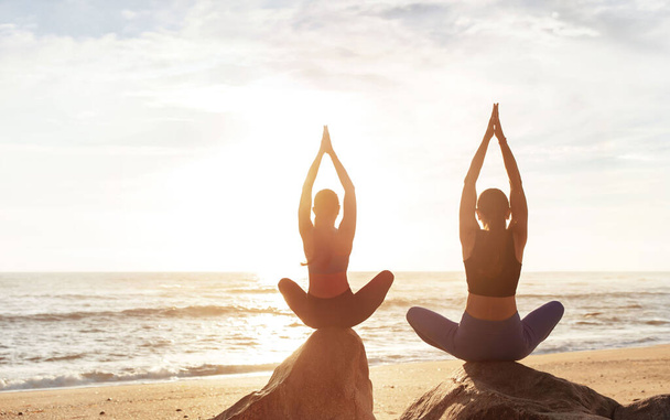 Feliz calma jovens mulheres caucasianas desfrutar de meditação, treino matinal, exercícios respiratórios, praticando ioga na posição de lótus na pedra, equilíbrio na praia do mar. Hábitos, estilo de vida e cuidados de saúde ao ar livre - Foto, Imagem