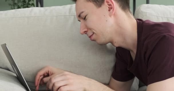 Iloisella tuulella, valkoihoinen mies makaa sohvalla ja innokkaasti tyypit hänen kannettava tietokone. Hänen iloinen luonteensa näkyy hänen kirkkaassa hymyssään, joka huokuu positiivisuutta ja onnea.. - Materiaali, video