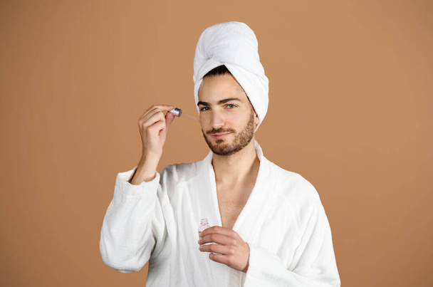 男性のスキン パンパリングと美容ケア. ホワイトバスローブナッシングスキンの中東男性,ベージュスタジオの背景にドロッパーを介してフェイシャルセラムを適用します. 男性のためのスキンケアのルートをリラックス - 写真・画像