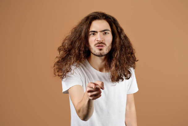 Портрет агресивного близькосхідного молодого хлопця з довгим волоссям вказуючи пальцем на камеру над бежевим тлом в студії. Негативні емоції, концепція токсичного спілкування. Вибірковий фокус - Фото, зображення