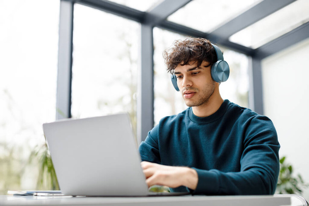 Молодой европейский дизайнер-мужчина слушает музыку на наушниках во время работы над ноутбуком за столом в современном стеклянном интерьере офиса, копировальное пространство. Фриланс, концепция онлайн-работы - Фото, изображение