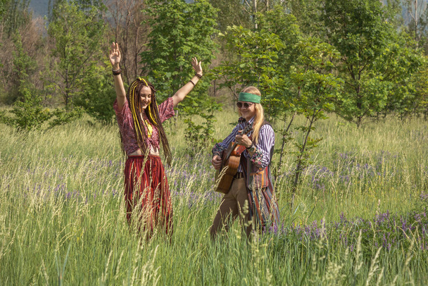 Couple of young people in hippie style. The girl is dancing, the guy is playing the guitar in a forest clearing on a sunny day - Photo, Image