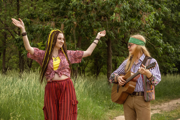 Hippi tarzı iki genç insan. Bir kız dans ediyor, bir erkek güneşli bir günde şehir yolunda gitar çalıyor. - Fotoğraf, Görsel