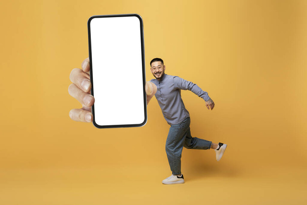 Funny Hombre asiático corriendo con enorme teléfono inteligente en blanco en la mano, alegre Millennial Guy Demostrando gran teléfono celular con pantalla blanca vacía, Posando sobre fondo de estudio amarillo, Collage, Mockup - Foto, Imagen