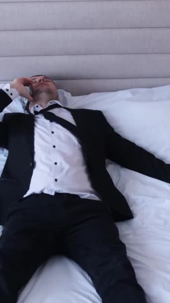 Un hombre cansado y relajado en un traje de negocios negro está acostado en una gran cama blanca y hablando por teléfono. Relajación después de un duro día de trabajo. Vídeo vertical - Metraje, vídeo