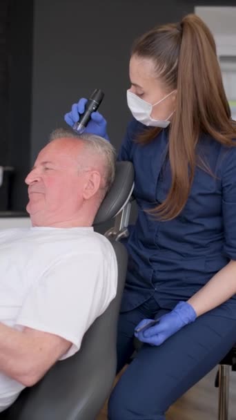 Um trichologist conduz um diagnóstico do cabelo e examina um homem mais velho. Conceito de tratamento capilar. Vídeo vertical - Filmagem, Vídeo