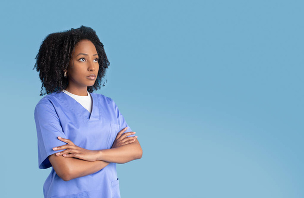 Smutna, poważna, czarna pielęgniarka w mundurze, skrzyżowana ramiona na klatce piersiowej, spogląda na wolną przestrzeń, odizolowana na tle niebieskiego studia. Problemy z egzaminem, pracą, medycyną i opieką zdrowotną - Zdjęcie, obraz