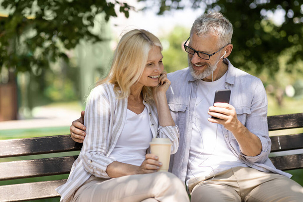 Romantic Older Couple With Smartphone Relaxing On Bench In Summer Park, Happy Mature Man And Woman Listening Music Together, Sharing Earphones And Drinking Takeaway Coffee, Enjoying Outdoor Leisure - 写真・画像
