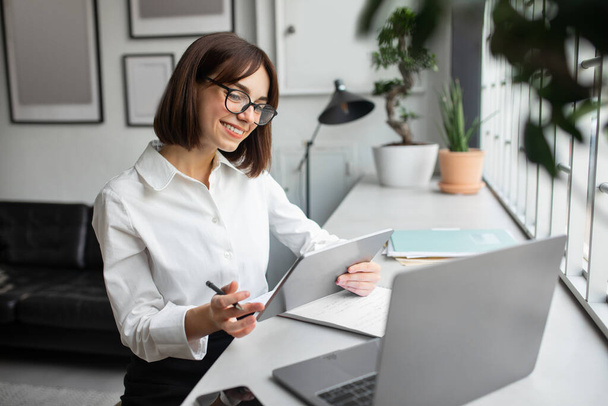 Pozytywna młoda bizneswoman pracująca z tabletem cyfrowym i laptopem, siedząca przy biurku w przytulnym biurze lub kawiarni, przedsiębiorczyni wykorzystująca nowoczesne technologie dla biznesu, korzystająca z networkingu - Zdjęcie, obraz