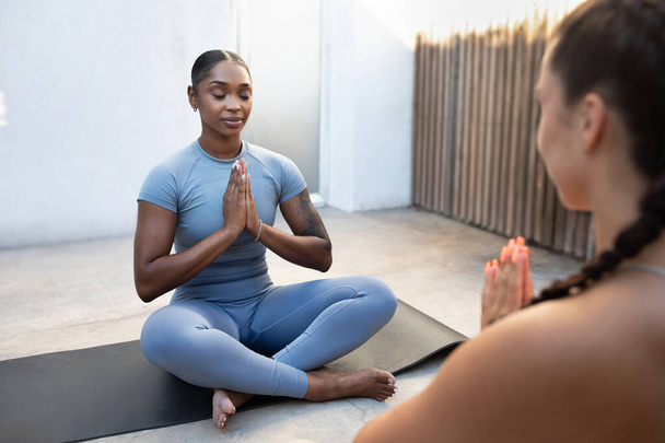 Calm smiling millennial black and caucasian women do breathing exercises in lotus position at morning, practice yoga together on mat, enjoy sport, workout, outdoor. Fit lifestyle, health care - Photo, Image