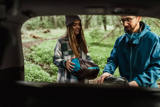 Uśmiechnięta młoda kaukaska para w kurtkach w lesie cieszy się wakacjami podróżnymi, bierze namiot z bagażnika, na świeżym powietrzu. Turystyka samochodowa, aktywny tryb życia, przygoda i turystyka, camping razem - Zdjęcie, obraz