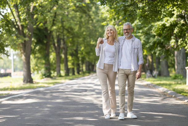 Portrait of beautiful senior couple walking in park together, full length shot of happy mature spouses relaxing outdoors, embracing and looking away, enjoying leisure after retirement, copy space - Photo, Image