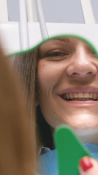 Frau bewundert Lächeln in Zahnarztpraxis. Glückliche junge Frau lächelt, während sie ihre perfekte Zahngesundheit im Spiegel in Großaufnahme überprüft. Vertikales Video - Filmmaterial, Video