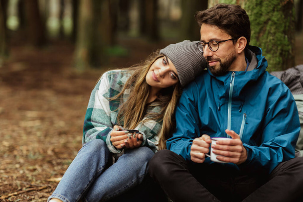 Relaxed happy young caucasian couple in jackets with cups of tea from thermos, enjoy travel, vacation, rest and hot coffee in forest, outdoors. Adventure and tourism, hiking, travel and love - Photo, Image