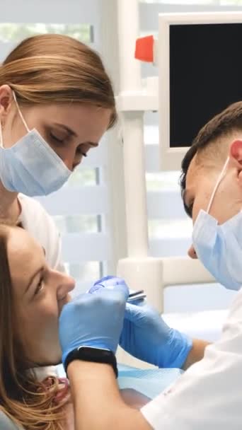 Egy fogorvos és egy asszisztens megvizsgálja egy női páciens fogait. Fogászati kezelés egy modern klinikán. Függőleges videó - Felvétel, videó