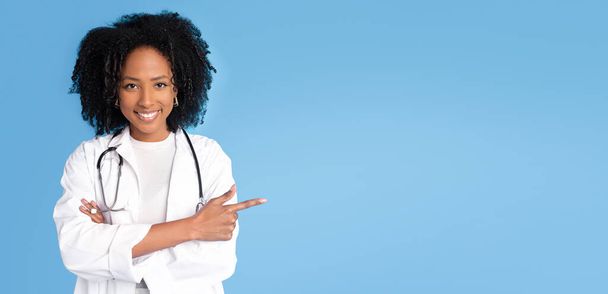 Médico mulher millennial preto positivo no dedo do ponto casaco branco no espaço de cópia, isolado no fundo do estúdio azul. Conselhos, recomendações, trabalho profissional do terapeuta, medicina e cuidados de saúde - Foto, Imagem