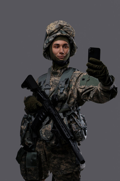 Nuori mies sotilasunivormussa ja kypärä kädessä kivääri ja älypuhelin, poseeraa selfielle tai ottaa videopuhelun harmaalla taustalla - Valokuva, kuva