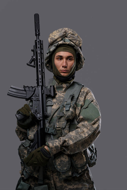 Молодой солдат в форме и шлеме позирует с серьезным выражением на сером фоне, демонстрируя силу и самоотверженность военнослужащих - Фото, изображение