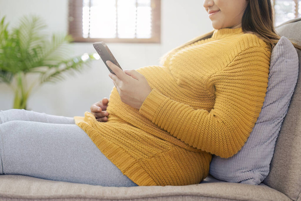Felice donna asiatica incinta utilizzando smart phone mentre si rilassa sul divano nel suo soggiorno. - Foto, immagini