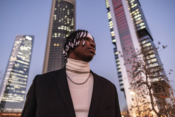 Low angle shot of an African American adult with braids in his hair, he is dressed in formal clothes and skyscrapers are seen in the background - Photo, Image