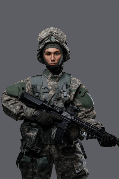 Молодой человек в военной форме и шлеме держит винтовку на сером фоне - Фото, изображение