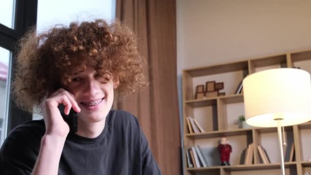Egy furcsa frizurájú fiatalember beszél telefonon. Boldog göndör vörös hajú fiatalember beszél a telefonban. Technológiák és kommunikáció. - Felvétel, videó