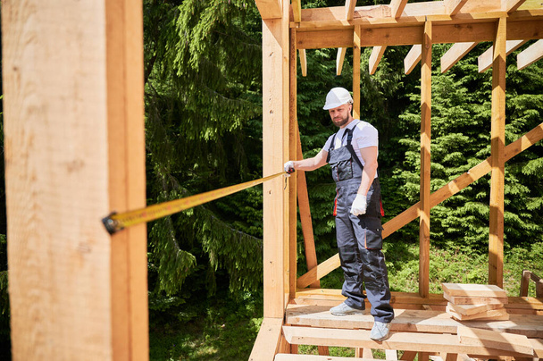 Carpenter constructing wooden skeleton building. Man measures distances with tape measure while wearing work attire and helmet. The concept of modern, environmentally-friendly construction. - Photo, Image