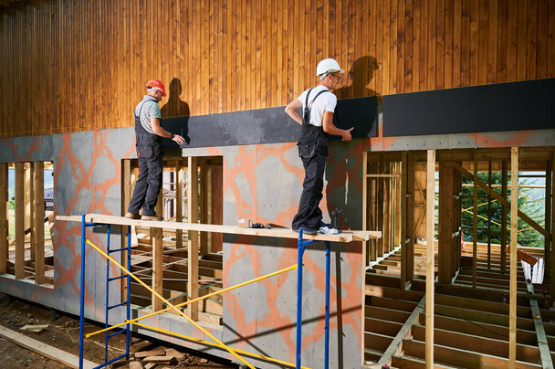 Asztalosok faházakat építenek. Két férfi munkás burkolja a ház homlokzatát cementlemezekkel, csavarhúzóval rögzítve őket. A modern környezetbarát építőipar koncepciója. - Fotó, kép