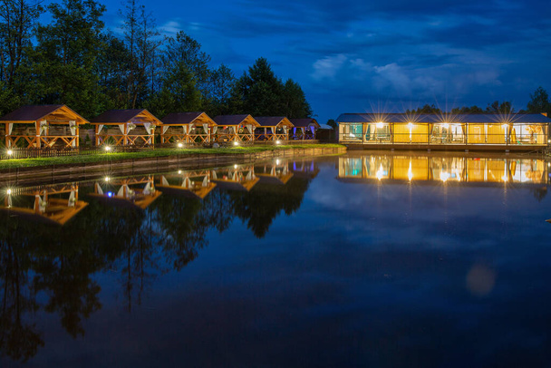 Fila de gazebos de madera para relajarse junto al lago y brillante pabellón o restaurante por la noche - Foto, imagen