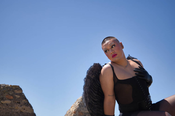 Portrait a non-binary person, young and South American, very makeup, with black wings and leather gloves, bodice and black stockings, sitting, posing next to the keep of a medieval castle. - Photo, Image