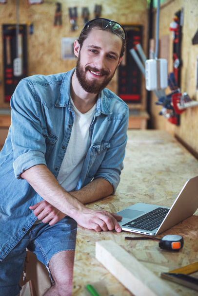 Bonito carpinteiro está usando um laptop, olhando para a câmera e sorrindo enquanto trabalhava na oficina
 - Foto, Imagem