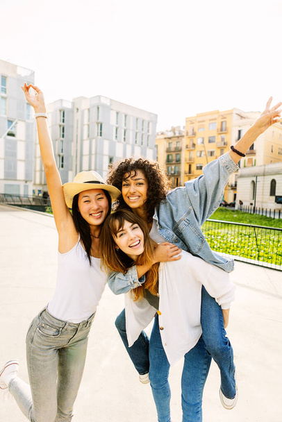 Νεαρή ομάδα τριών πολυφυλετικών γυναικών διασκεδάζουν απολαμβάνοντας καλοκαιρινές διακοπές στο δρόμο της πόλης. Γυναικεία ιδέα φιλίας με διαφορετικές έφηβες κοπέλες να γελάνε μαζί έξω. - Φωτογραφία, εικόνα
