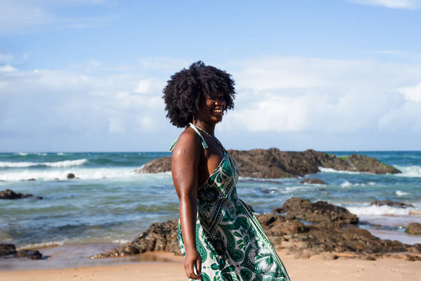 Портрет красивой темноволосой женщины, улыбающейся и счастливой стоя на пляже. На заднем плане облака скал и моря. Рио-Вермелью, Сальвадор, Бразилия. - Фото, изображение