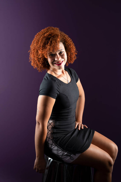 Portrait of a beautiful attractive woman with red hair, sitting, smiling and happy, posing for a photo in black clothing against a lilac background. - Photo, Image