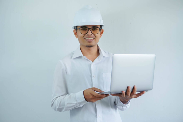 Junger asiatischer Architekt trägt Bauarbeiter-Schutzhelm über isoliertem Hintergrund und freut sich, Laptop in der Hand zu halten. - Foto, Bild