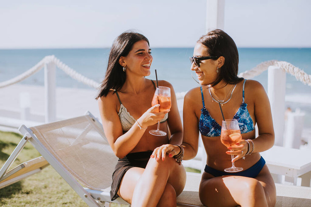 カクテルを飲んでビーチで休暇を楽しんでいるビキニの2人の笑顔の若い女性 - 写真・画像