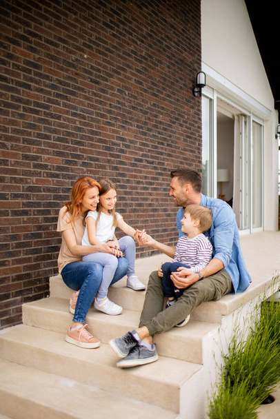 Сім'я з матір'ю, батьком, сином і дочкою, що сидить надворі на сходах переднього ганку цегляного будинку
 - Фото, зображення