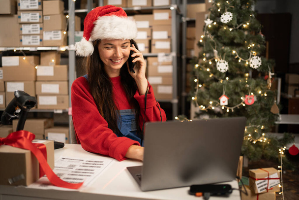Warehouse manager in Santa hat pick up phone purchase order stock detail customer online data delivery at storehouse. Startup small business concept. - Photo, Image
