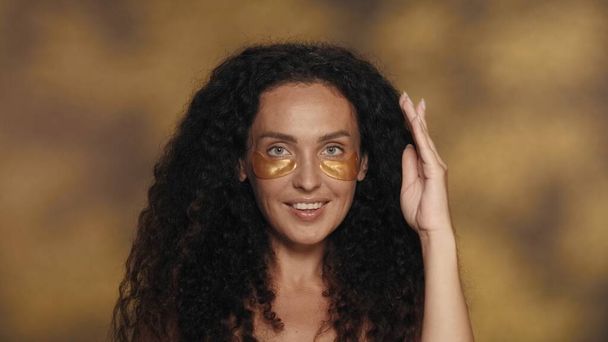 Longhaired curly brunette woman performs cosmetic procedures. Portrait of a seminude woman with golden patches under her eyes in the studio on a yellow background with highlights close up. The concept - Zdjęcie, obraz