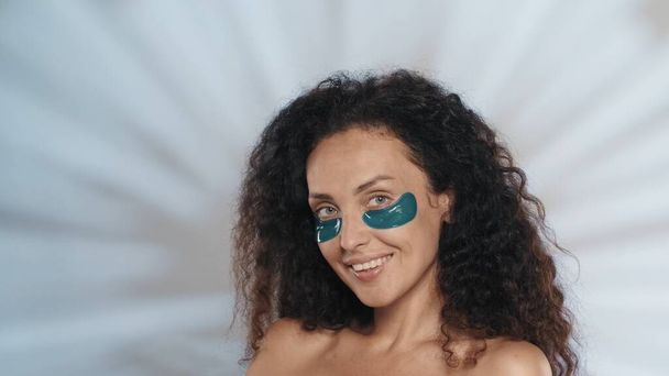 Une femme effectue une procédure cosmétique. Portrait d'une femme séminude avec des taches bleues sous les yeux dans le studio sur un fond bleu avec des reflets. Beauté, cosmétologie, soin de la peau - Photo, image