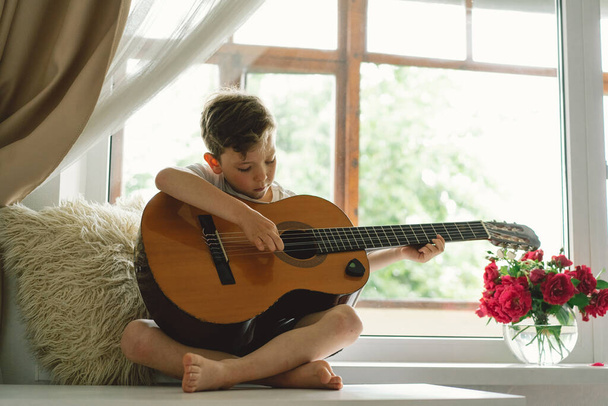 Lindo chico aprende a tocar la guitarra clásica en el alféizar de la ventana cerca de la ventana. Acogedora casa. Vacaciones de verano estilo. - Foto, imagen