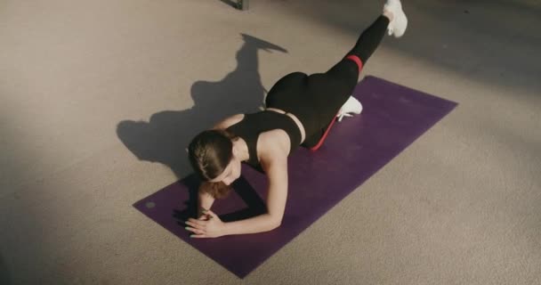 Sportowa brunetka ćwicząca w odzieży sportowej, zostaje na desce z paskiem oporu i podnosi nogę. Sportowa kobieta trenuje na macie do ćwiczeń na świeżym powietrzu - Materiał filmowy, wideo