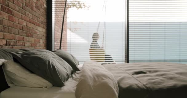 Fechar na cama confortável vazio com almofadas e cobertor coberto no quarto moderno com interior loft e pessoa balançando fora atrás janela panorâmica com persianas, câmera lenta - Filmagem, Vídeo