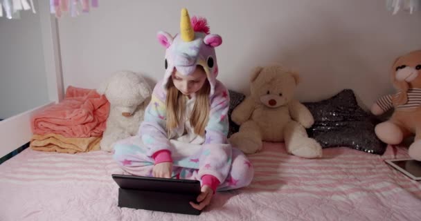 The 7-year-old girl is playing in the childrens room in unicorn costume and using digital computer tablet, enjoying cool video or photo content in social network, playing online games. - Materiaali, video