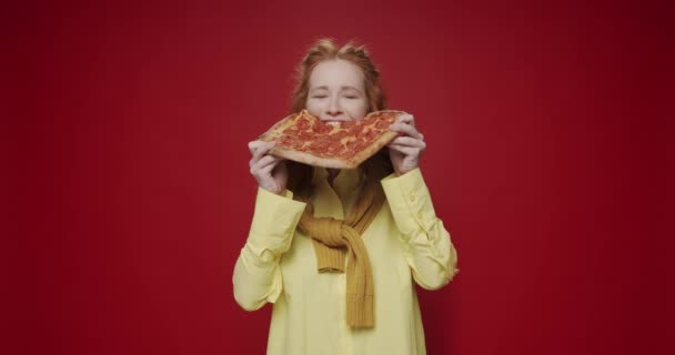 Het mooie hongerige meisje dat peperoni pizza eet, geniet en glimlacht op een rode geïsoleerde achtergrond. Hongerige vrouw geniet van Pizza. Een lekker stukje Pepperoni pizza eten. - Video