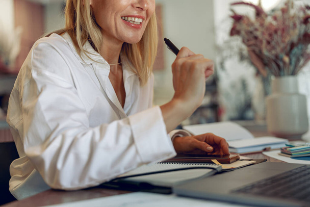 Усміхнена жінка-підприємець працює на ноутбуці, роблячи нотатки в затишному інтер'єрі коворкінгу - Фото, зображення