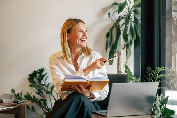 Uśmiechnięta kobieta przedsiębiorca pracuje na laptopie, notatki w przytulnym współpracownikiem wnętrza przestrzeni - Zdjęcie, obraz